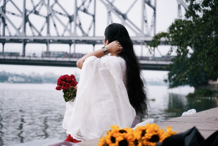 A woman sitting by a bridge gazing with flowers in hand