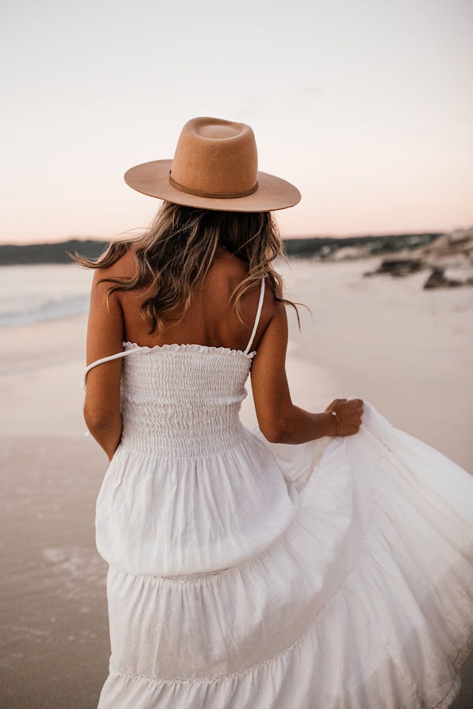 A woman in a white dress and hat walks along a serene beach at sunset. Reinvent yourself after 35 and start building a life that truly