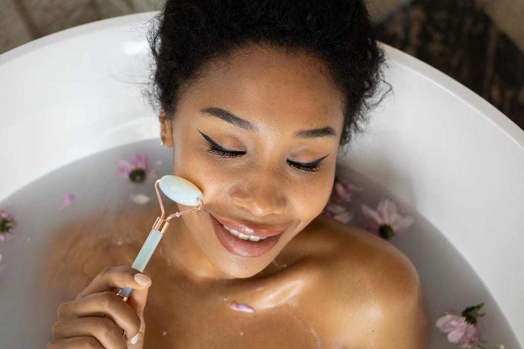 From above of smiling ethnic female with closed eyes massaging face with jade roller while taking bath with flowers Skincare for Women Over 35: Glow, Protect, and Breathe