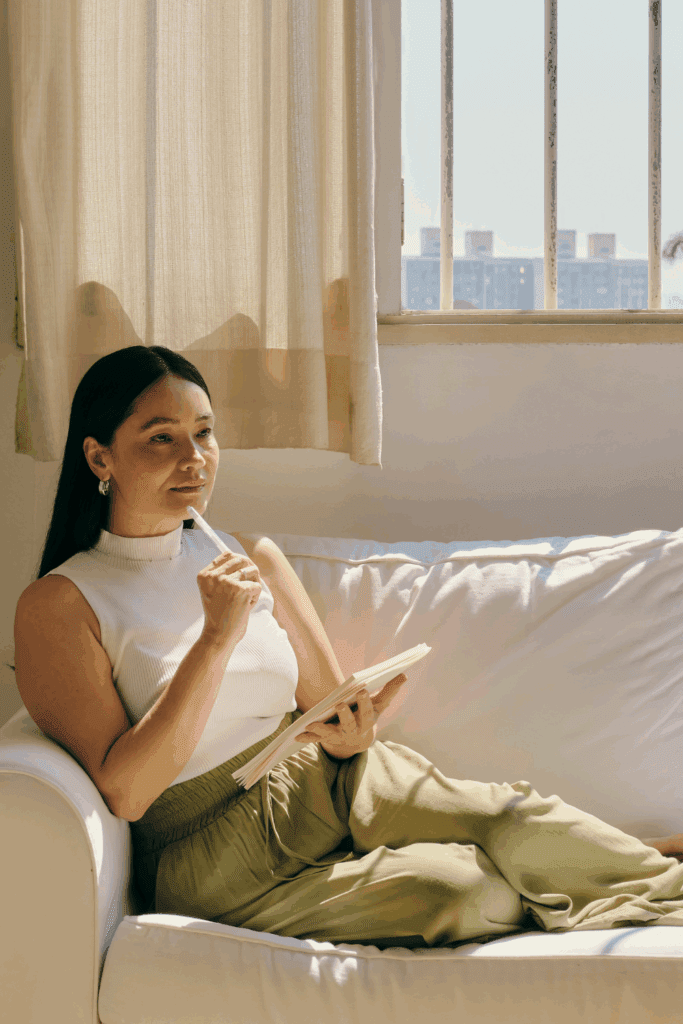 A woman in her late 30s to 50s journaling by a window, soft morning light.