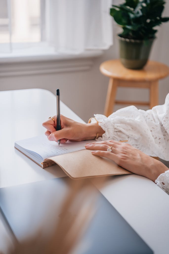 Close-up of a woman writing in a JOURNAL at a bright home office desk.