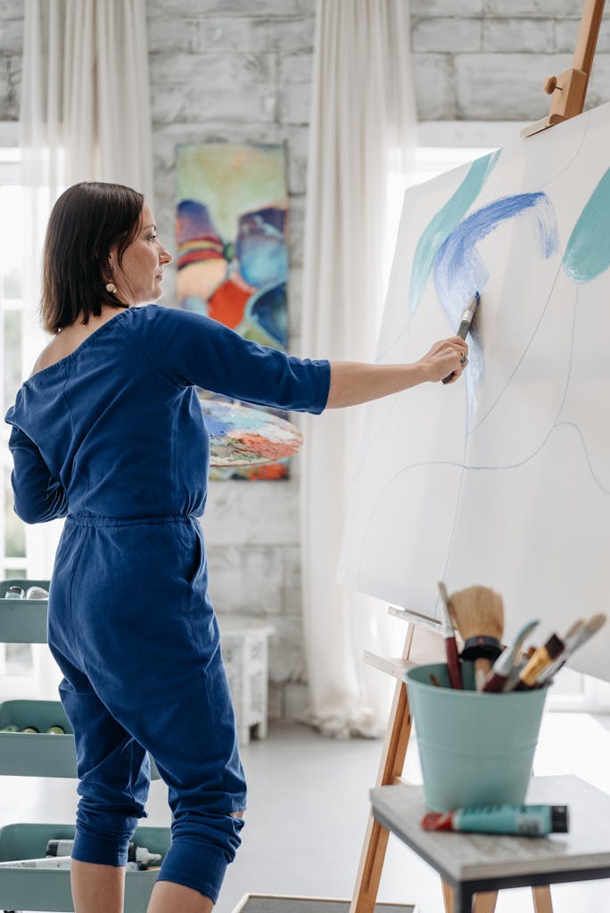 Woman artist in blue jumpsuit painting a colorful abstract on large canvas indoors.