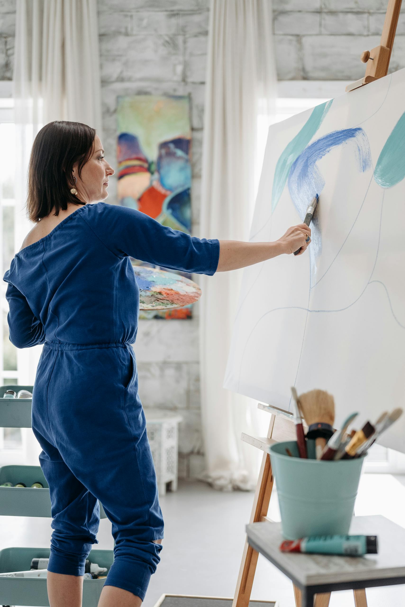 Woman artist in blue jumpsuit painting a colorful abstract on large canvas indoors.
